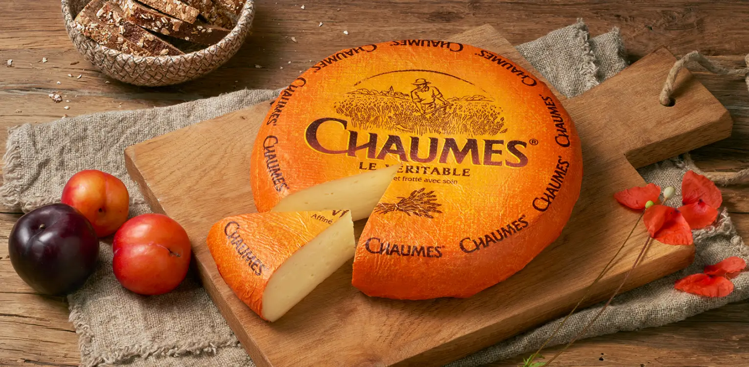 Chaumes cheese wheel with one wedge cut out, displayed on wooden board with apples, flowers, and bread basket on rustic wooden surface.