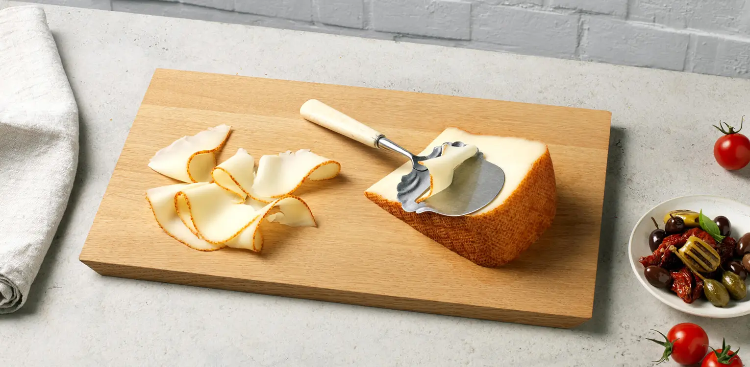 Wooden cutting board with apple peels, a peeler tool, and a halved apple, surrounded by tomatoes and a bowl of vegetables on white surface.
