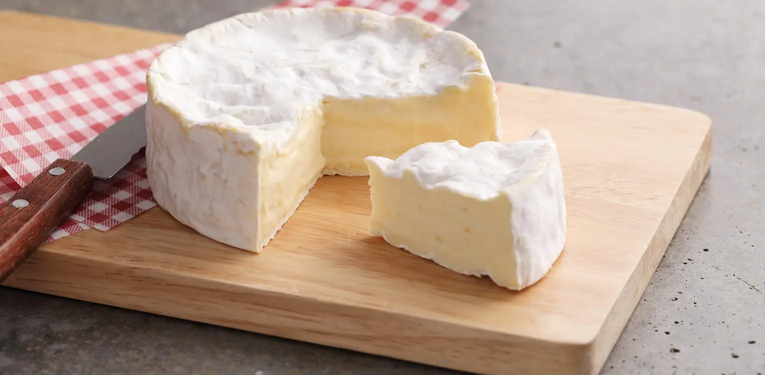 Soft ripened cheese with white rind on wooden board, one slice cut away, red checkered cloth beside it.