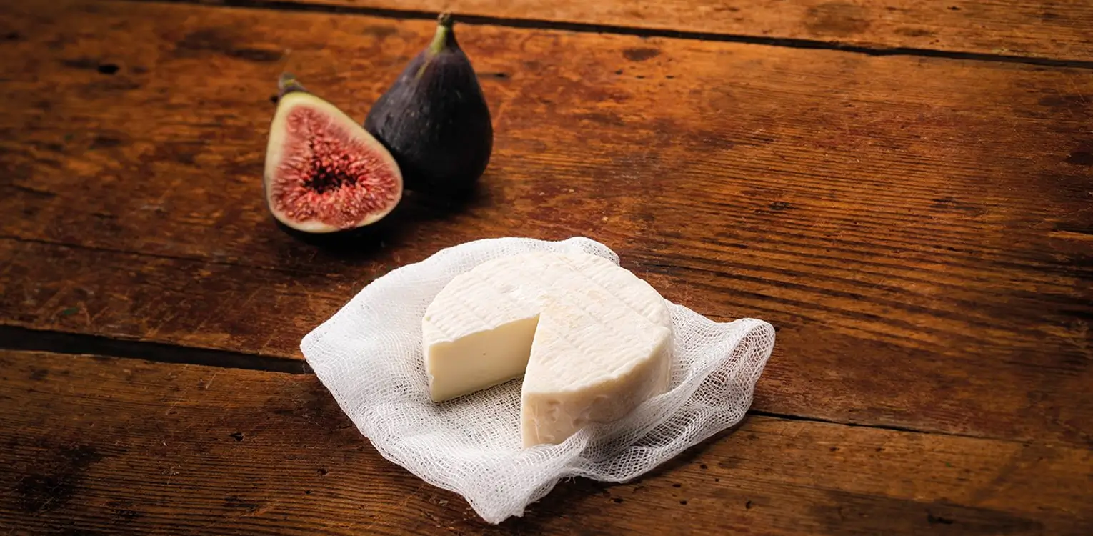 Two wedges of fresh white cheese on parchment paper beside a halved fig with dark skin and pink interior on wooden surface.
