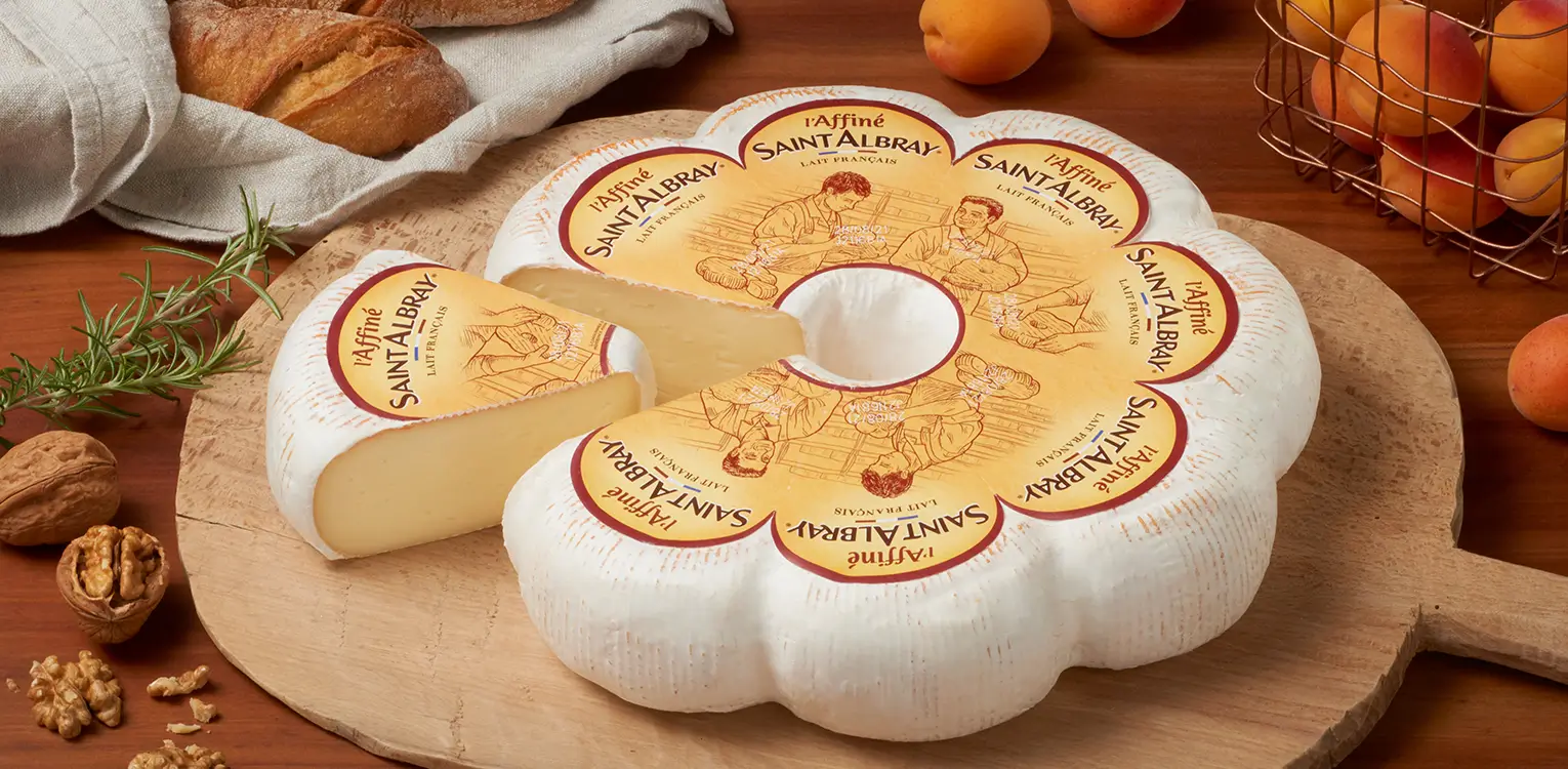 Flower-shaped Saint-Albray cheese wheel with decorative label on wooden board, surrounded by walnuts, bread, and fresh apricots.