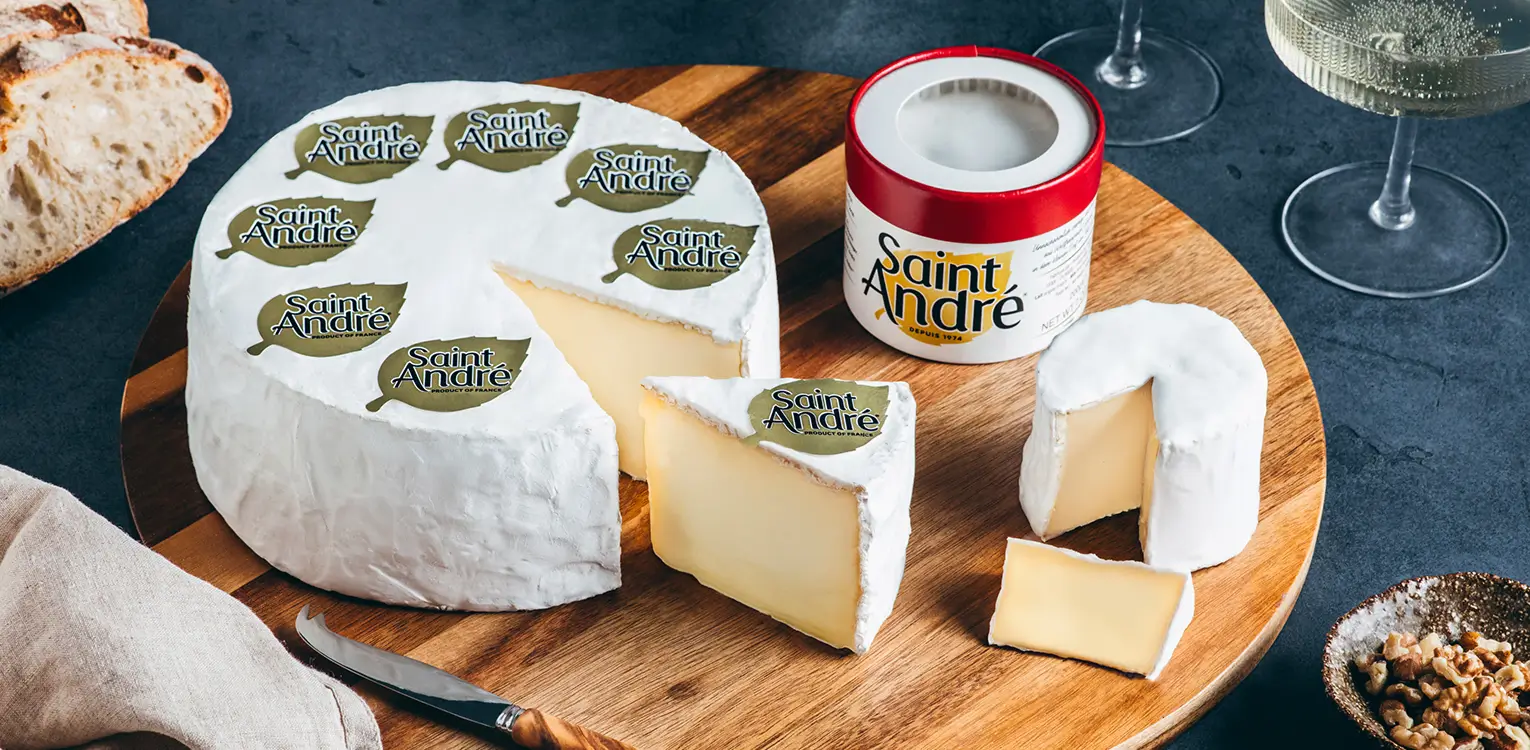 Saint André cheese display on wooden board showing whole wheel with white rind, cut slices, and branded packaging jar with wine glasses and bread.