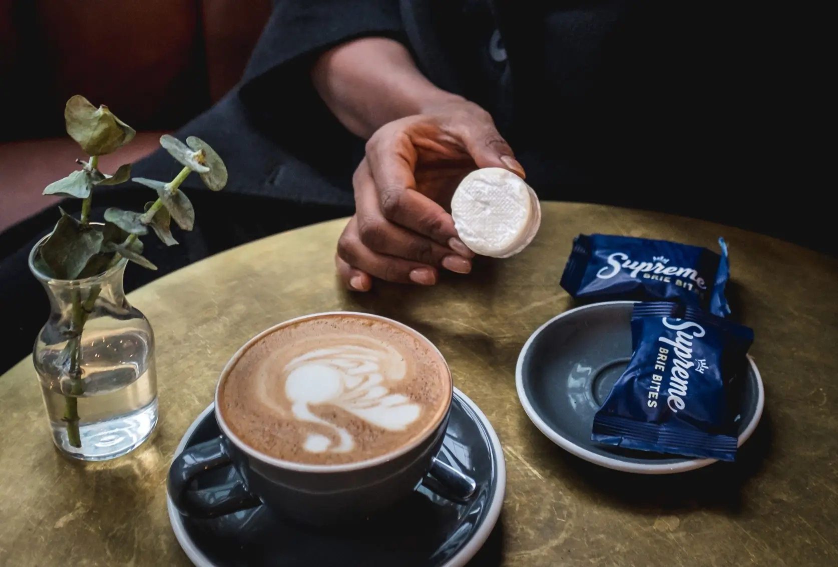 Person holding a sugar packet above a latte with latte art, beside a small vase with greenery and sugar packets on a gold table.