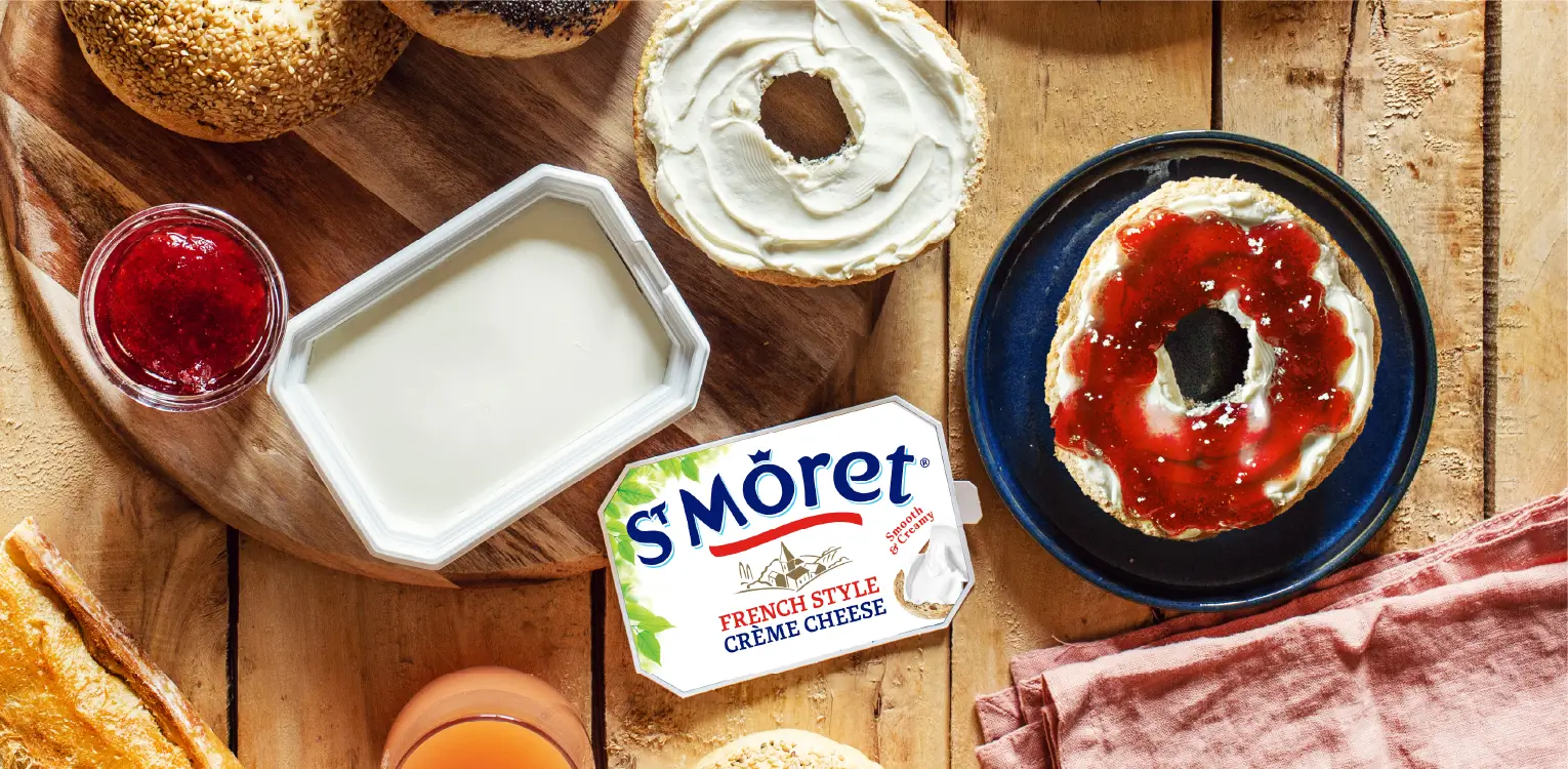 Overhead view of bagel assembly ingredients on wood surface: Smorét cream cheese package, fresh cream cheese in bowls, jam in glass, sliced bagels, and cured meat on cloth.
